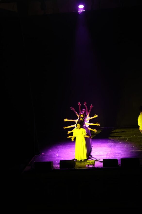 Group of young dancers in yellow dresses standing in a line with arms outstretched under a purple spotlight on stage.