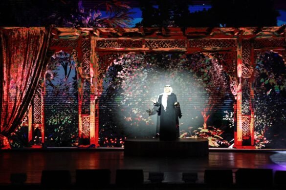 Performer in a black robe and white headscarf stands on a lit round platform, with a red ornate garden backdrop behind.