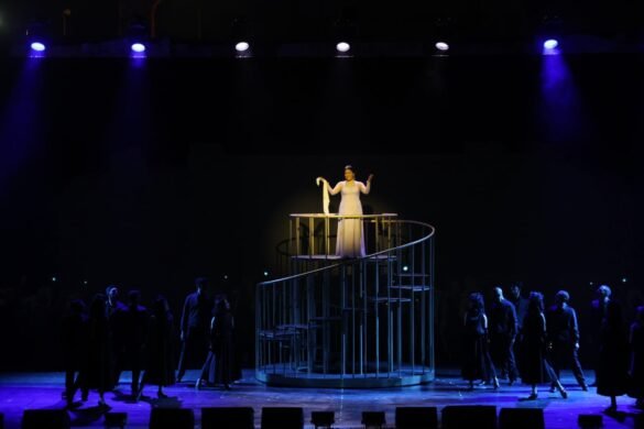 A performer in a light gown stands on a spiral metal staircase center stage, holding a cloth, under bright spotlights while a dark audience watches.