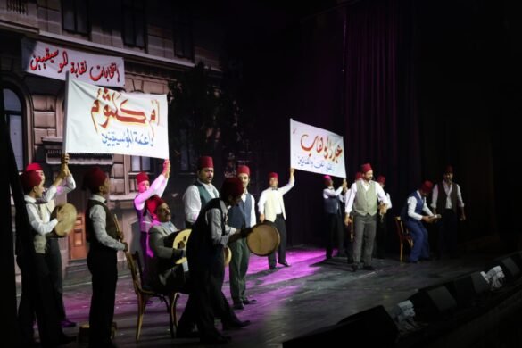Group of men in fez hats and vests perform on a stage, some playing drums while others hold banners with Arabic text.