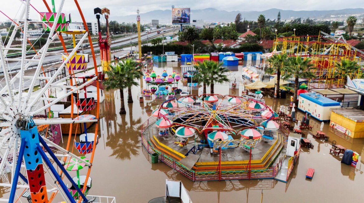 Aerial view of a colorful amusement park flooded with brown water, showing a Ferris wheel, rides, and palm trees from above.