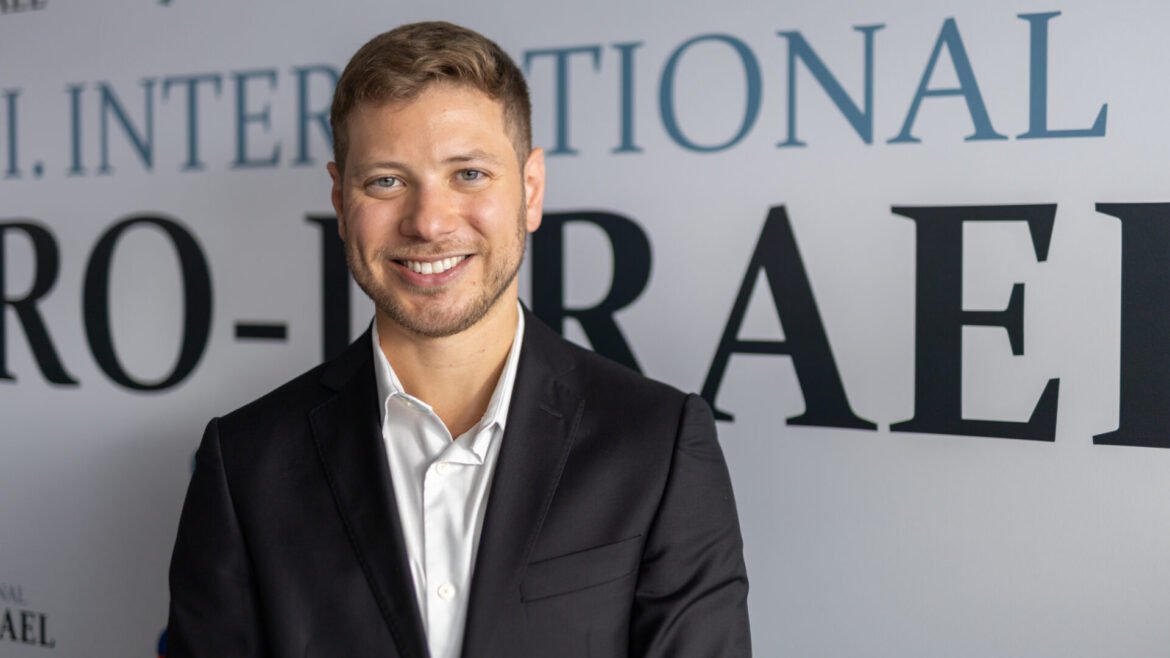 Smiling man in a dark suit and white shirt posing for a professional portrait in front of a wall with large black letters.