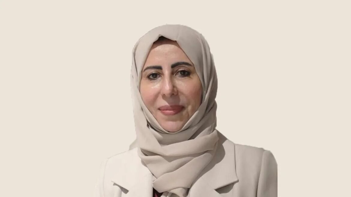 Portrait of a woman wearing a beige hijab and light blazer, smiling softly against a neutral tan background.