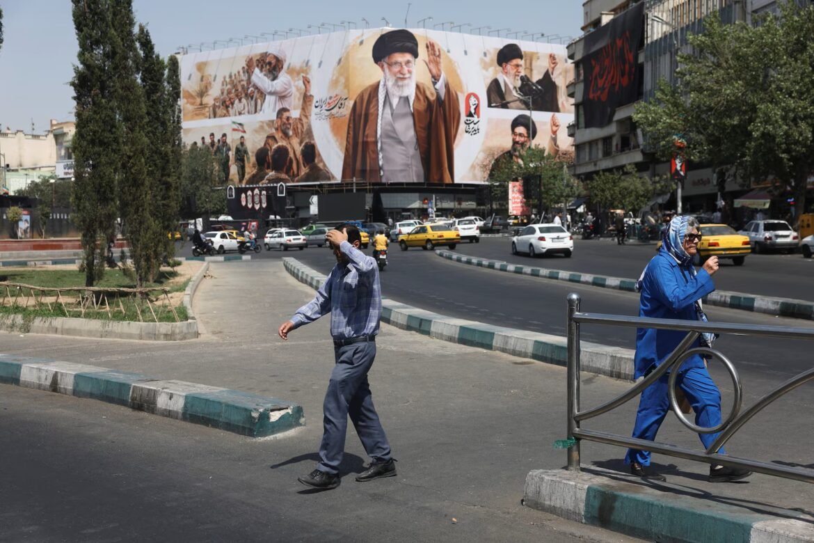 City street with a giant mural of Iranian leaders on a building; pedestrians cross the road in the foreground.