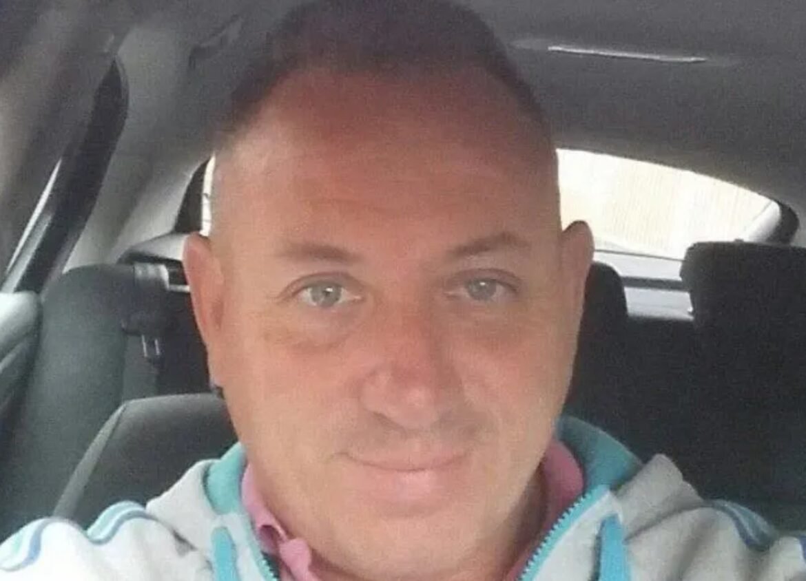 Selfie of a man sitting in a car, wearing a light hoodie with teal accents, looking at the camera.
