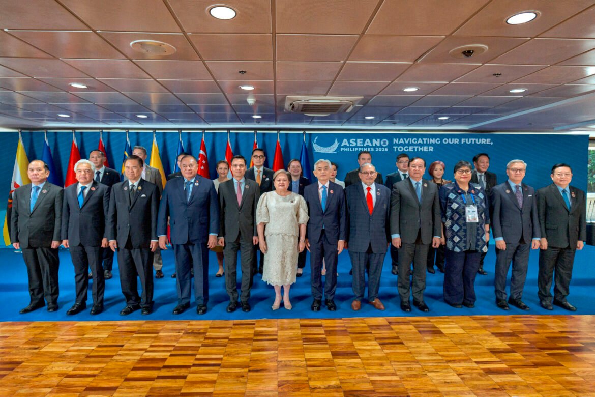 Group of officials in formal attire posing for a photo on a stage with ASEAN flags and a blue backdrop plate saying ASEAN 2026.