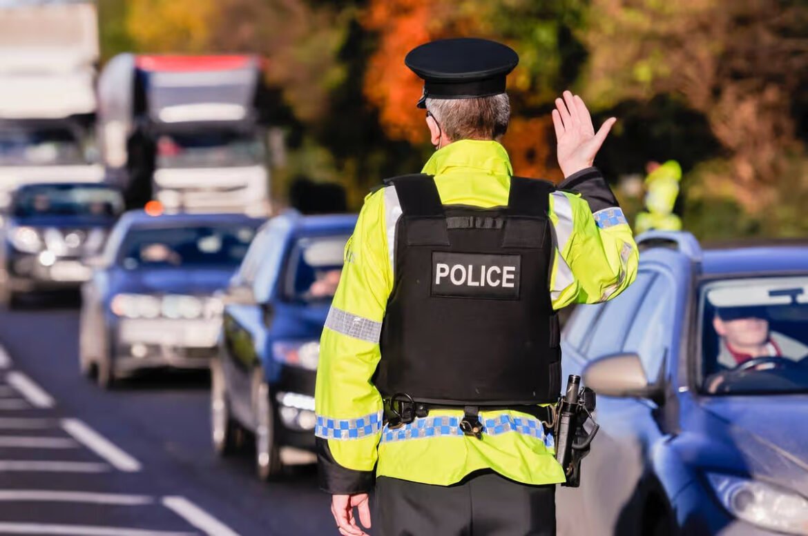 Police officer in a bright jacket standing on a road, facing traffic and waving to cars.