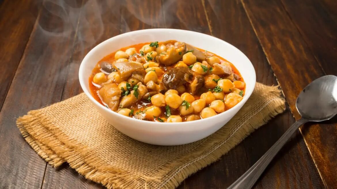 Chickpea and mushroom stew in tomato sauce, garnished with parsley, served in a white bowl on a wooden table with a burlap mat and steam rising up.