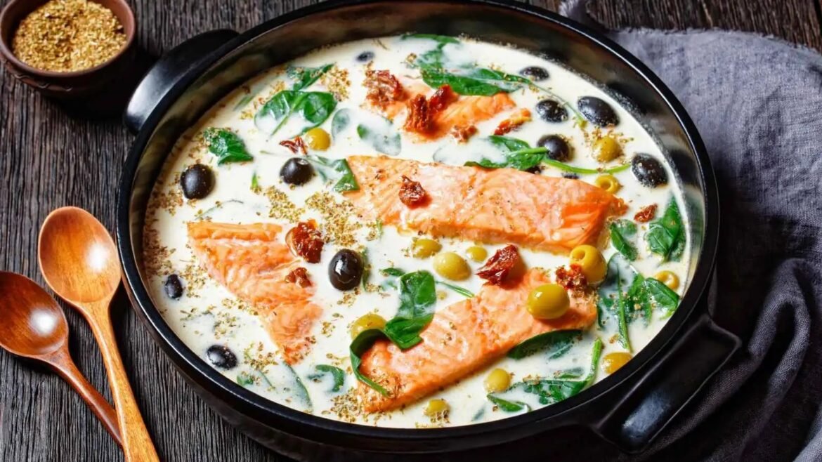 Salmon fillets simmering in a creamy white sauce with olives, spinach, and herbs in a black skillet.
