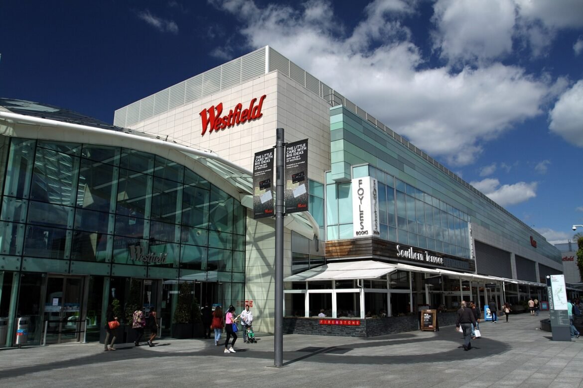 Westfield shopping centre exterior with glass entrance and a large red Westfield sign against a blue sky.