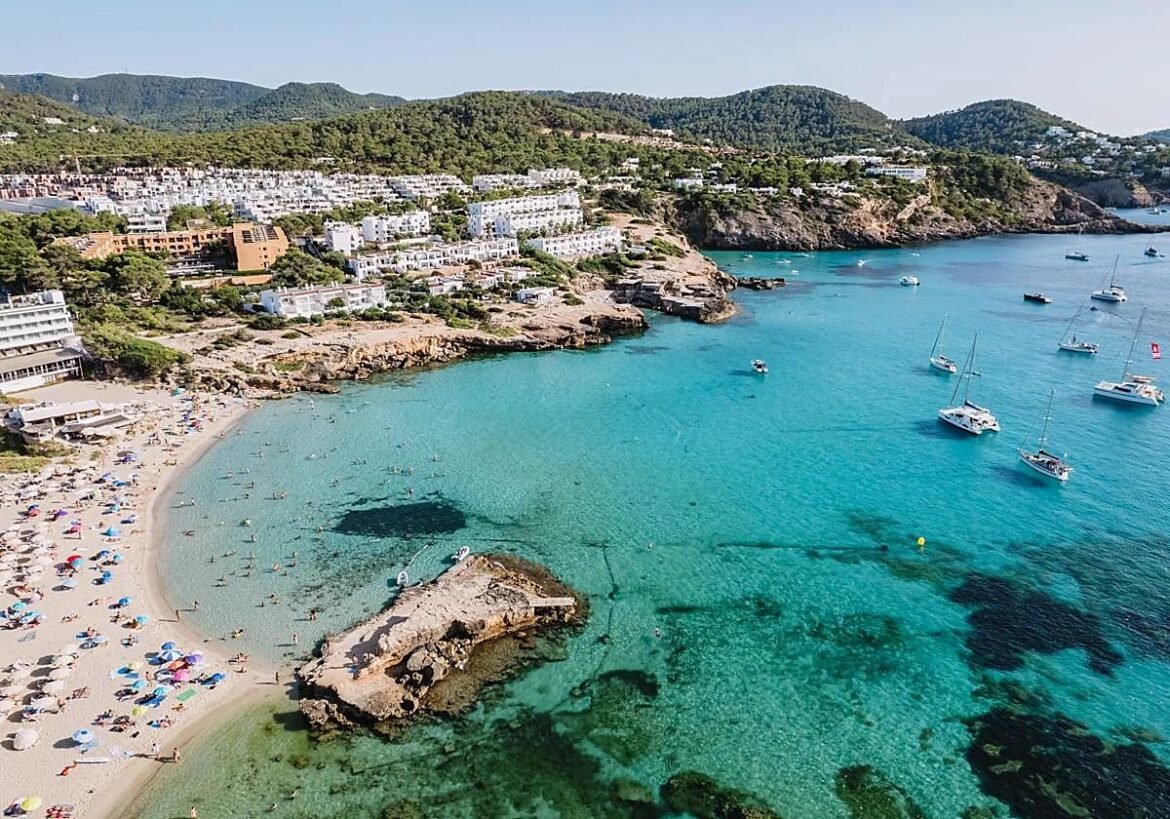 Aerial view of a seaside town with white buildings on green hills, a rocky promontory, and a turquoise bay with boats.
