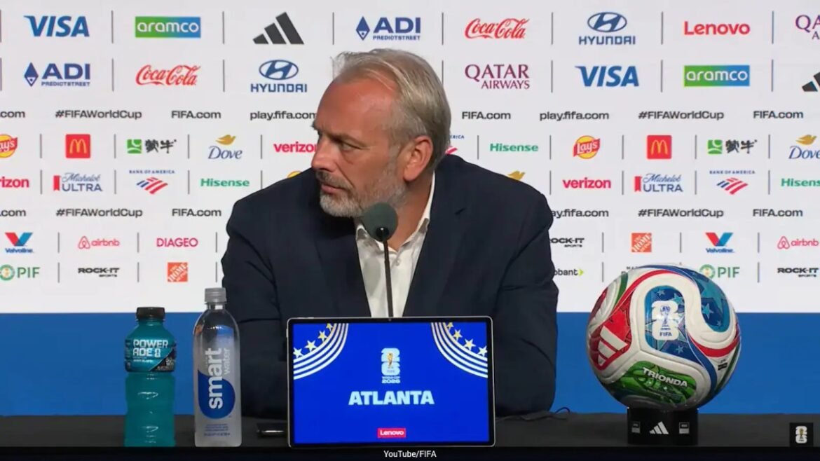 Man in a dark suit at a press conference table with a microphone; laptop screen reads ATLANTA with FIFA World Cup graphics; a soccer ball on a stand to the right, and water bottles nearby.
