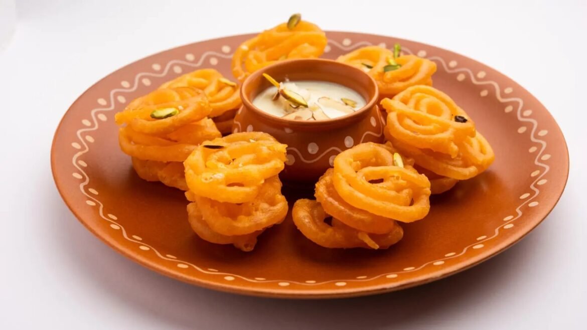 Plate of orange jalebi spirals arranged around a small bowl of creamy dipping sauce, with almond slices atop the dip.