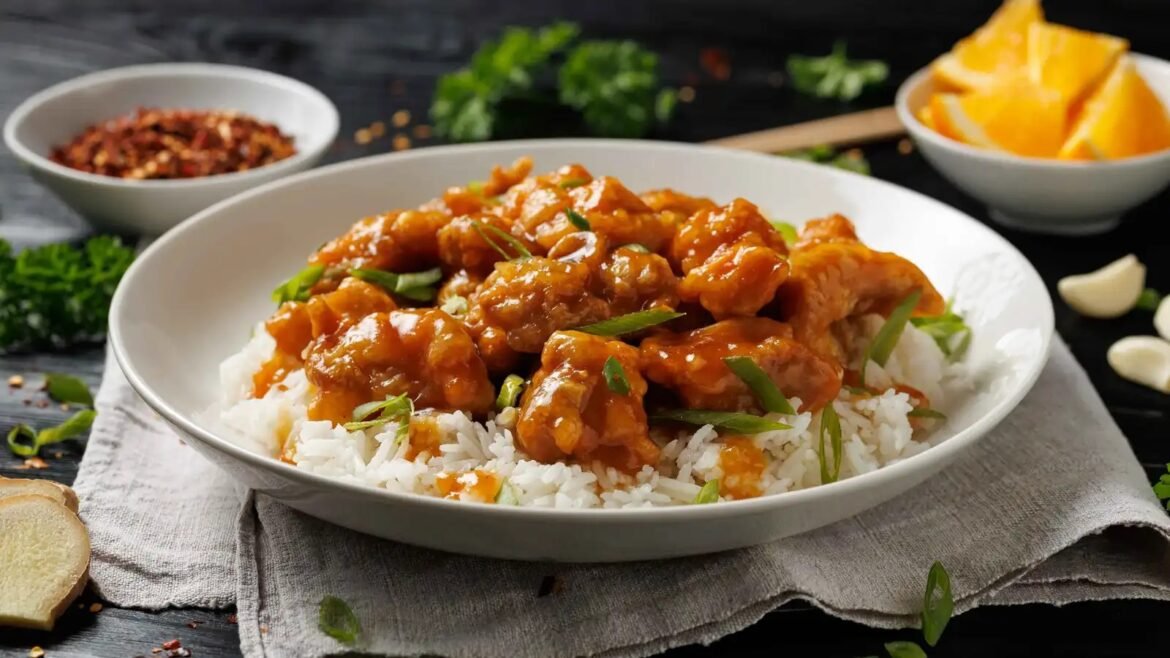 Bowl of white rice topped with orange glazed chicken and chopped green onions on a gray napkin. Background bowls and garlic cloves create a cozy dining scene.