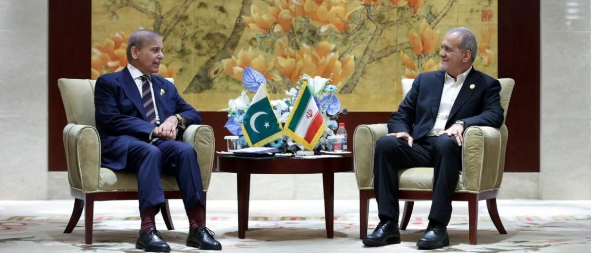 Two male diplomats in suits sit in beige chairs facing each other, with Pakistan and Iran flags on a table between them.