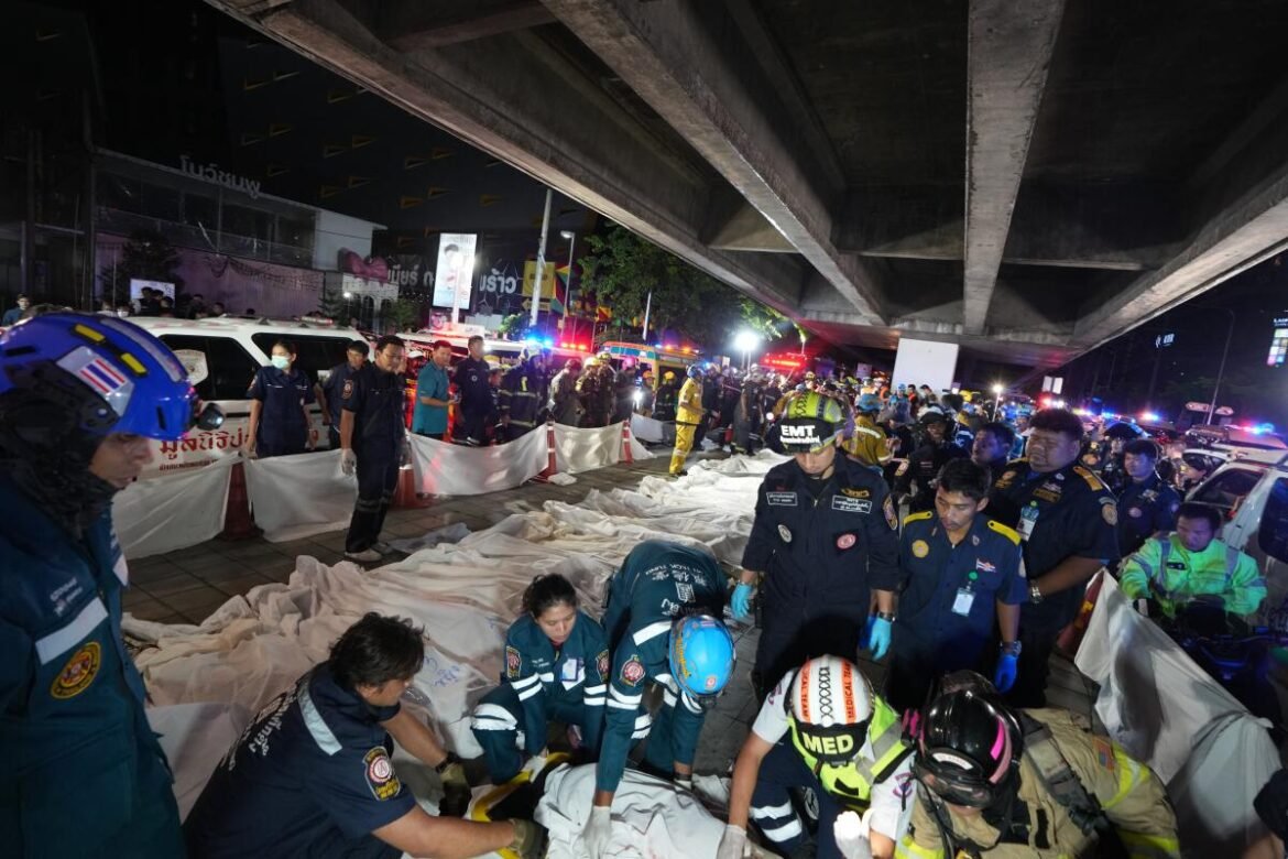 Emergency responders in uniforms work around white body bags under a concrete roadway at night, with police and ambulances in the background under an overpass.