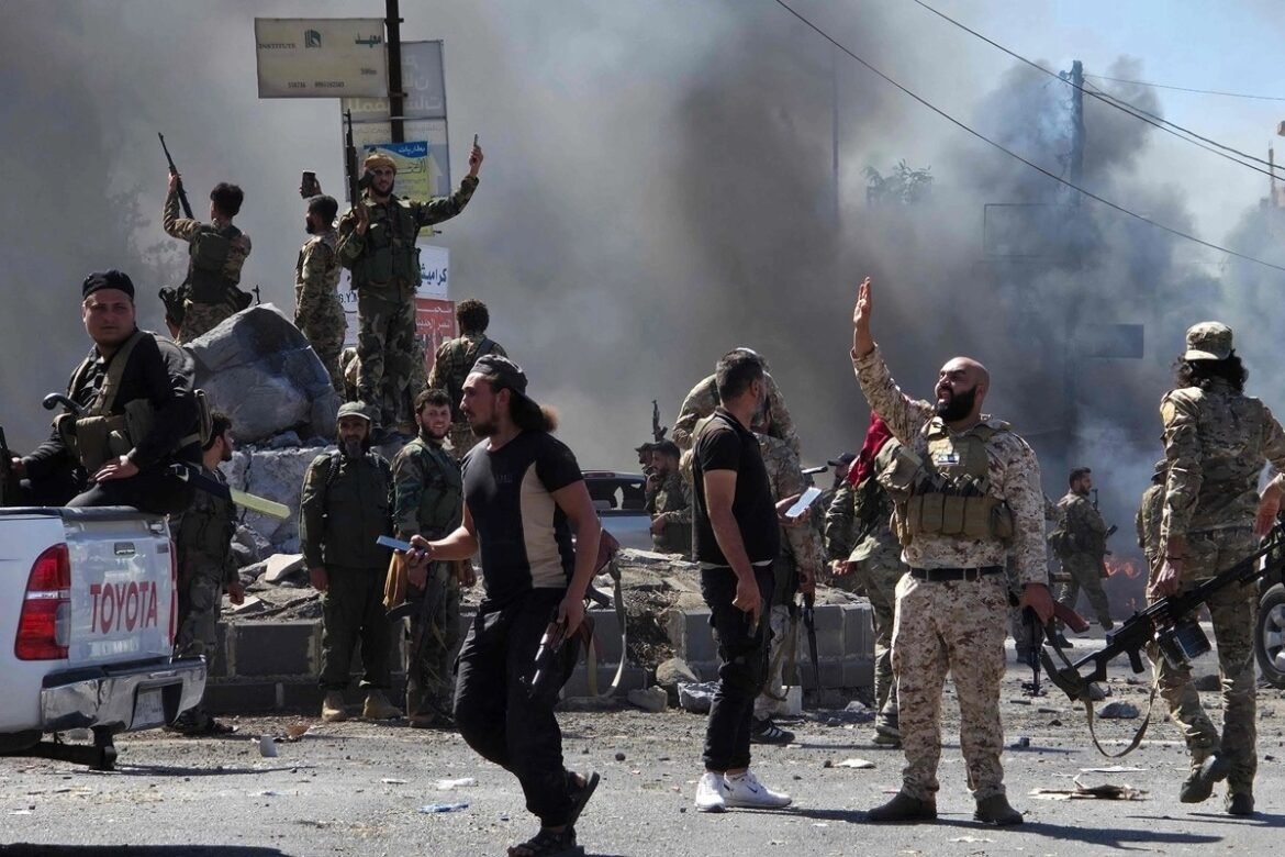 Militants in camouflage and desert uniforms gesture and carry rifles amid smoke and rubble on a damaged street, with a white Toyota pickup in the foreground and debris around them as some stand on a pile.