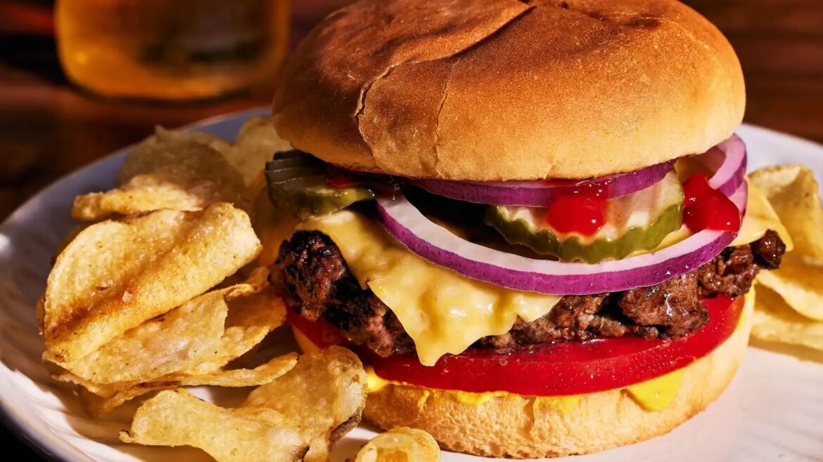Juicy cheeseburger with melted cheese, pickles, red onion, tomato, and mustard on a bun, served with potato chips.