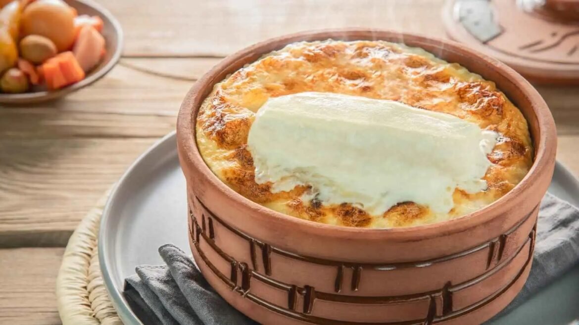 Baked dish in a clay pot with a melted cheese topping and a pale cheese slab on top, served on a wooden table.