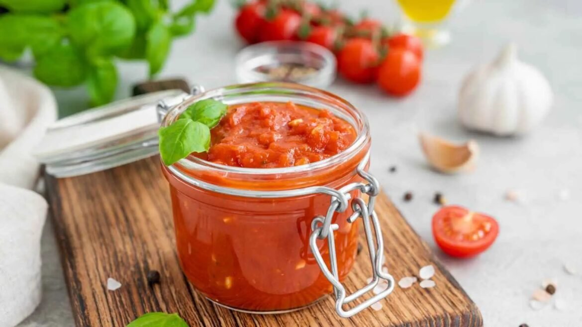 Glass jar of chunky tomato sauce with a basil garnish on a wooden board, with tomatoes and garlic in the background