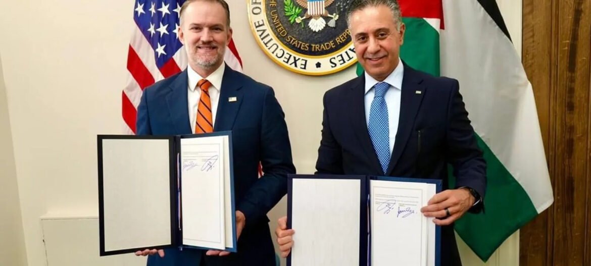 Two men in suits hold open folders with signed documents during a formal signing ceremony, with a government seal and flags behind them.