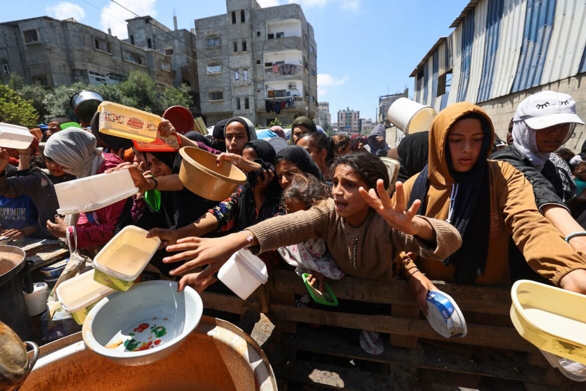 Crowd of people, mainly women, reach for bowls during a relief food distribution in a dense urban area with unfinished buildings nearby.