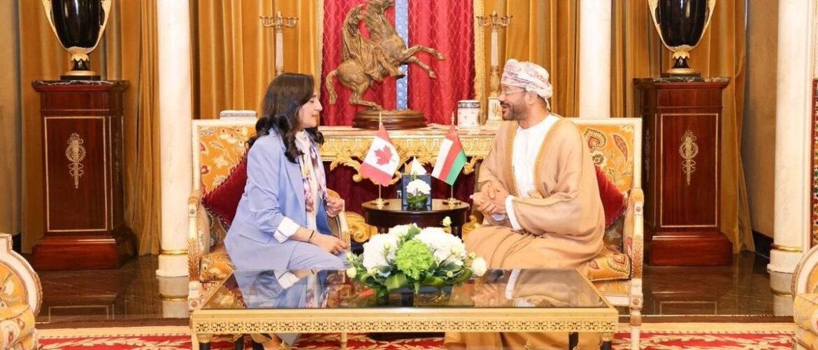 Two people sit cross‑legged on an ornate sofa in a lavish room, a small table with Canadian and UAE flags between them as they chat.