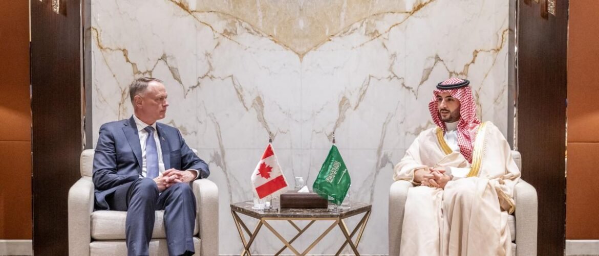 Two men sit in light chairs across from each other in a formal meeting, with Canadian and Saudi flags on a small table between them.