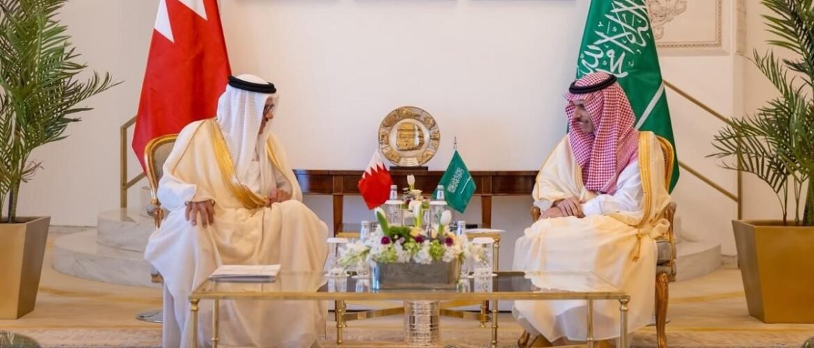 Two men in white thobes sit across from each other in a formal room, with Bahrain and Saudi flags behind them and a flower centerpiece on the table.
