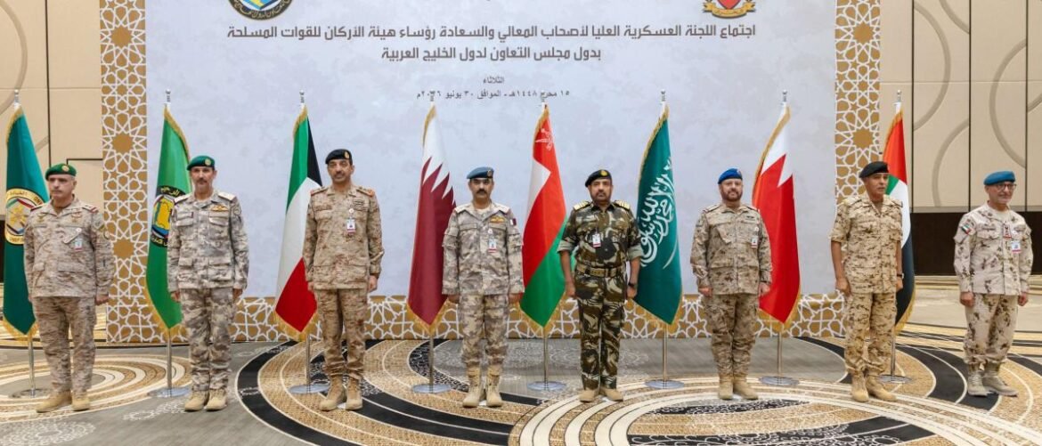 Nine military officers in desert camouflage stand in a row with national and organizational flags behind them at a formal ceremony.