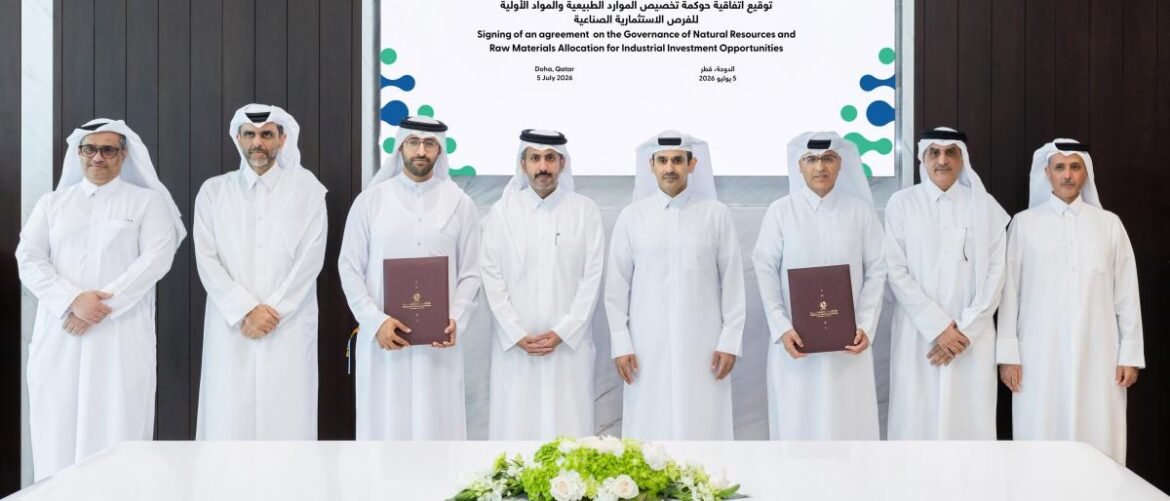 Group of men in white kanduras stand in a row at a signing ceremony, two holding brown folders in front of a banner screen.