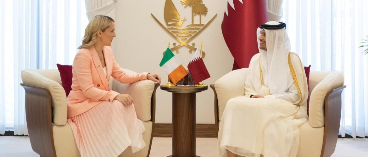 Diplomatic meeting between a woman in a peach suit and a man in traditional white attire, seated with Ireland and Qatar flags on a small table between them
