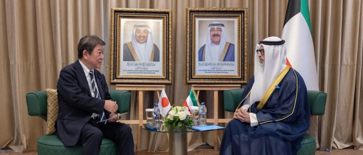 Two men in formal attire sit facing each other in a diplomatic meeting, with Japan and Kuwait flags on the table and framed portraits behind them.