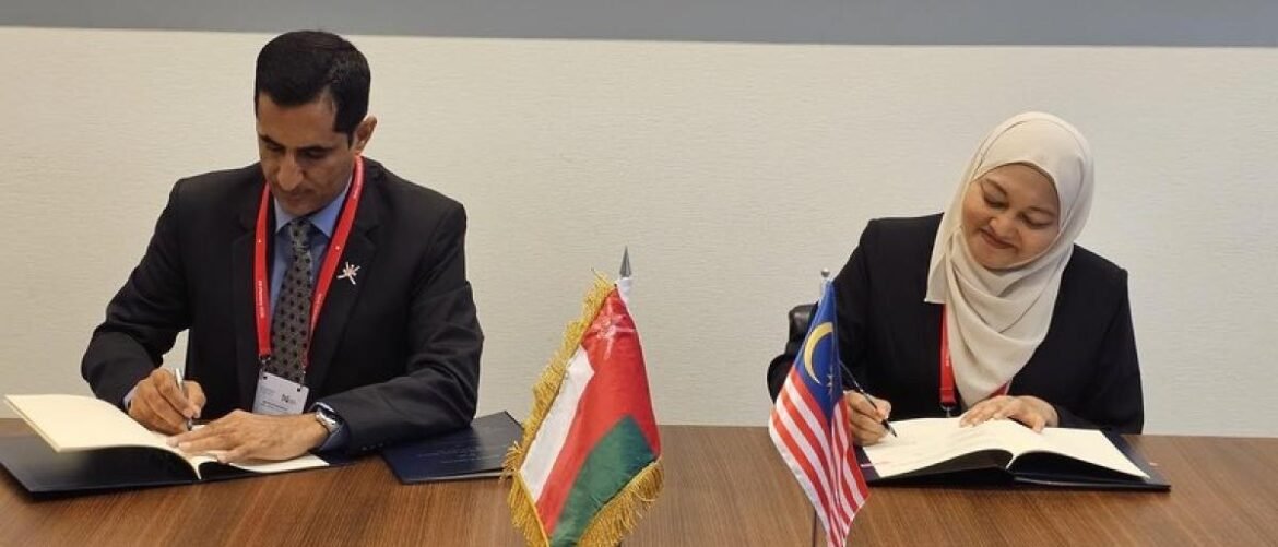 Two professionals sign documents at a conference table, with the Oman and Malaysia flags between them on the table in front of a formal setting.