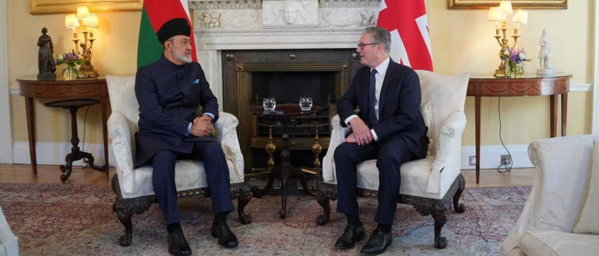 Two men in suits sit in white armchairs facing each other in a formal room with flags and a marble fireplace behind them.
