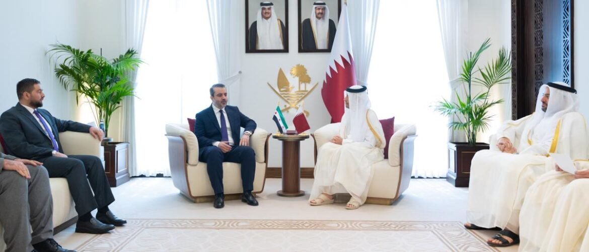 Diplomatic meeting in a bright room with the Qatari flag and national emblem in the background; five men seated on sofas around a small table.