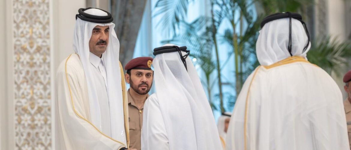 Two men in white thobes and head coverings greet each other indoors, with a security officer in uniform watching in the background.