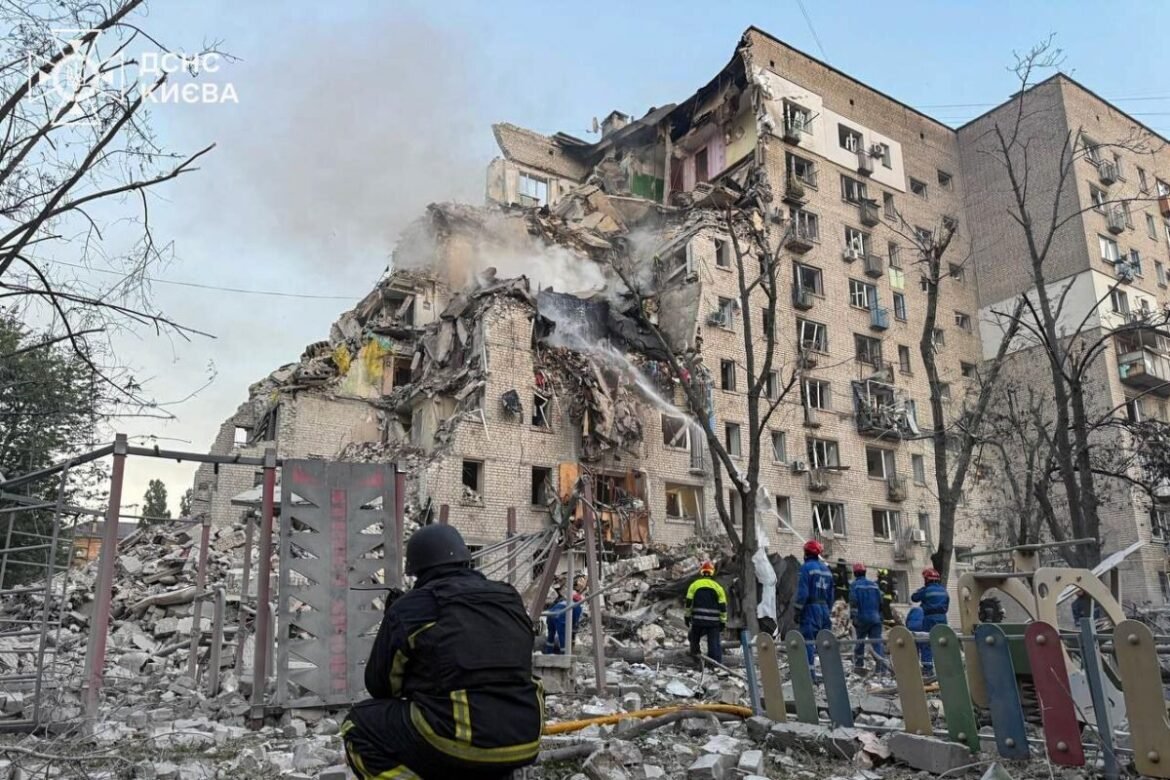 Collapsed multi-story apartment building with smoke, rescuers in helmets and reflective vests at the rubble.