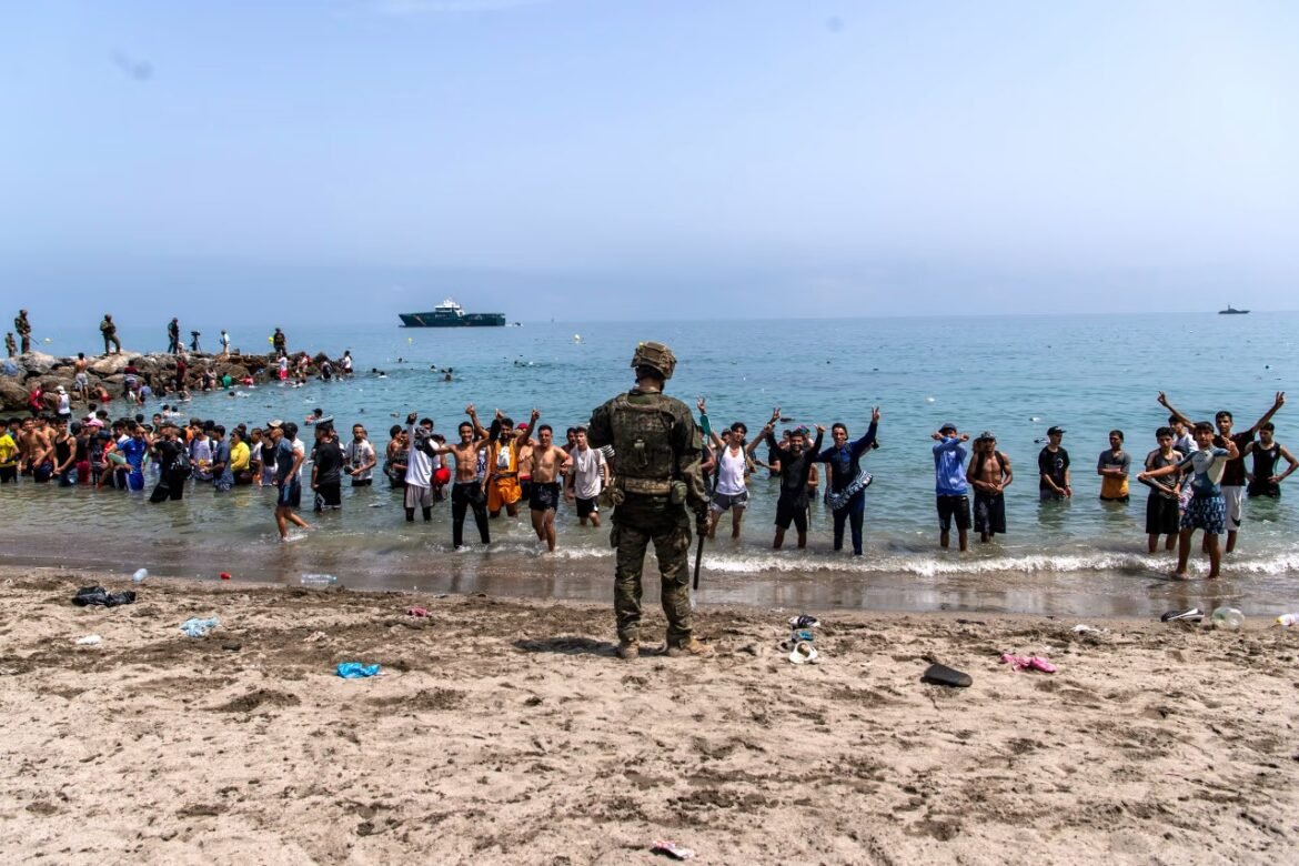 Soldier in camouflage stands on a sandy beach facing a group of people waist-deep in the sea, some raising their hands, with a ship in the distance.
