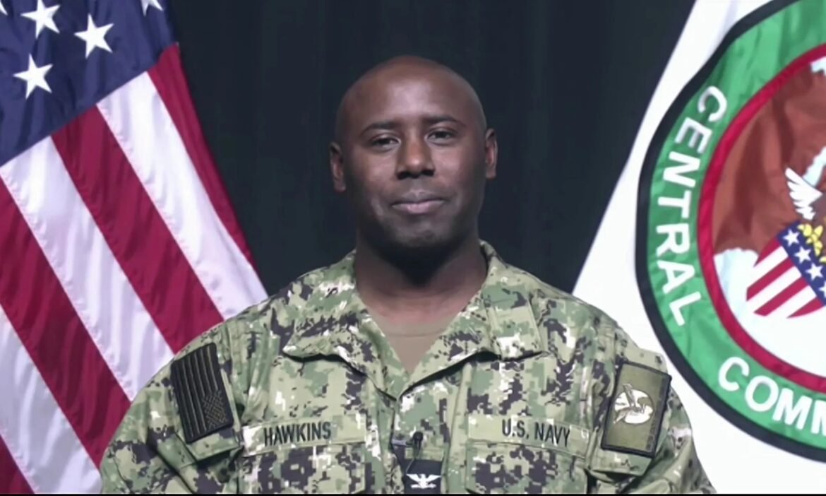 Portrait of a Black U.S. Navy sailor in camouflage uniform named Hawkins, standing in front of the American flag and a ceremonial seal backdrop.