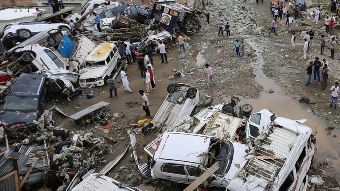 Crowd of people standing among a chaotic pile of crushed and overturned vehicles and rubble at a muddy disaster site.