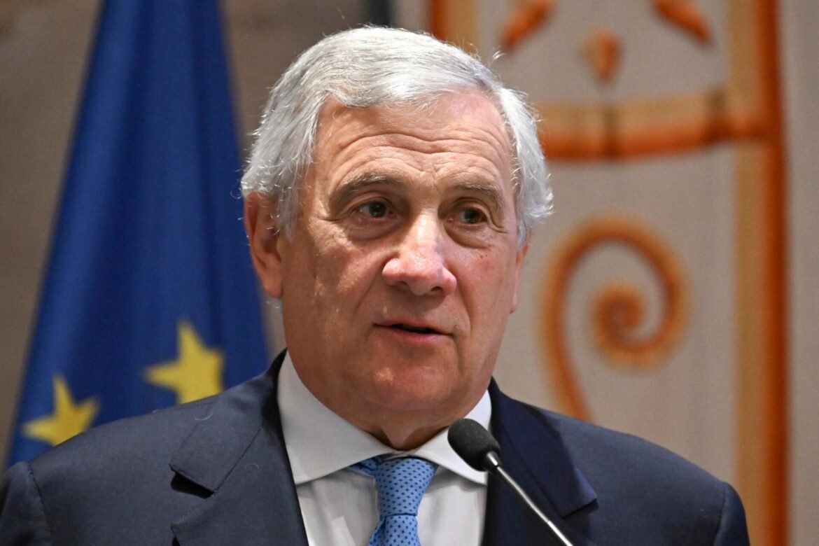 Close-up of a middle-aged man in a dark suit and blue tie speaking into a microphone, with a European flag in the background.