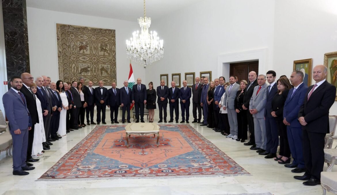 Group of men and women in formal attire standing along the walls of a grand room with a chandelier and a Lebanese flag in the background.