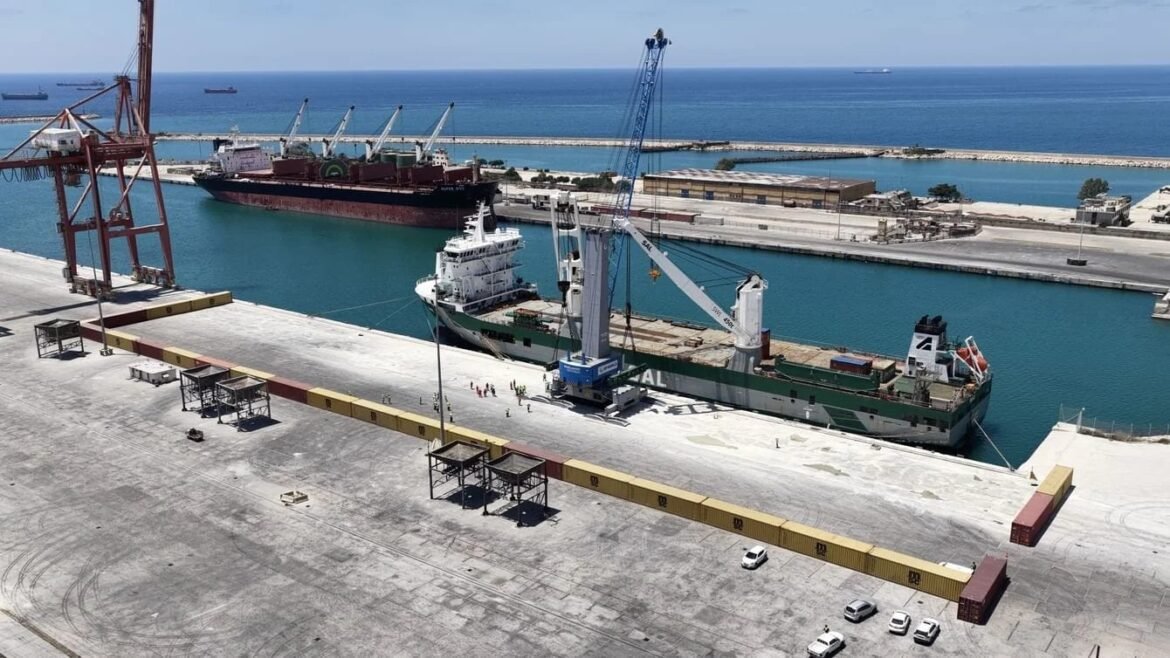 Aerial view of a busy port: a large cargo ship moored at the dock with cranes overhead and a crane barge nearby, stacks of containers along the quay, and several cars parked on the concrete foreground with clear blue water beyond.