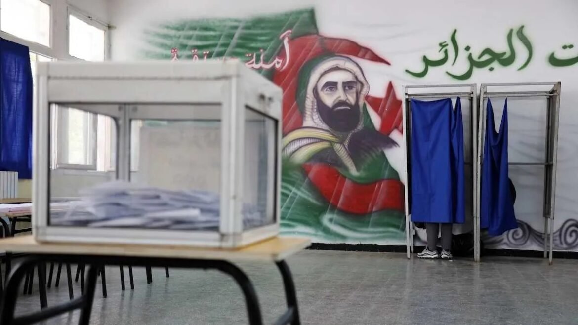 Polling station with a transparent ballot box on a table and blue privacy booths against a wall mural in the background.