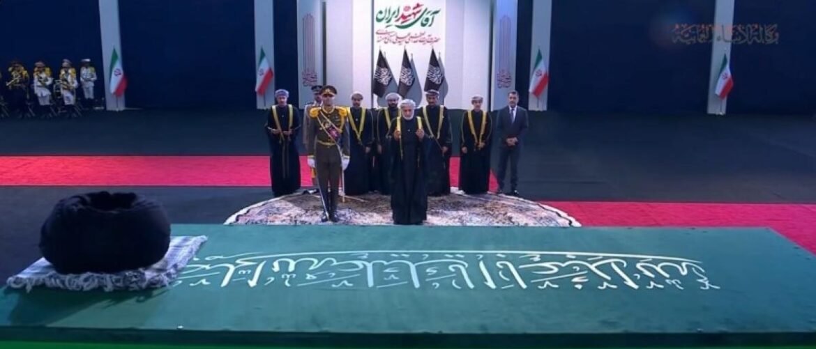 Group of officials in ceremonial robes stand on a stage with Iranian flags; foreground shows a green table with white script.