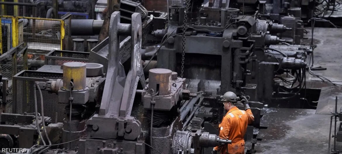 Worker in orange safety suit and hard hat operating a large metal press on an industrial factory floor.