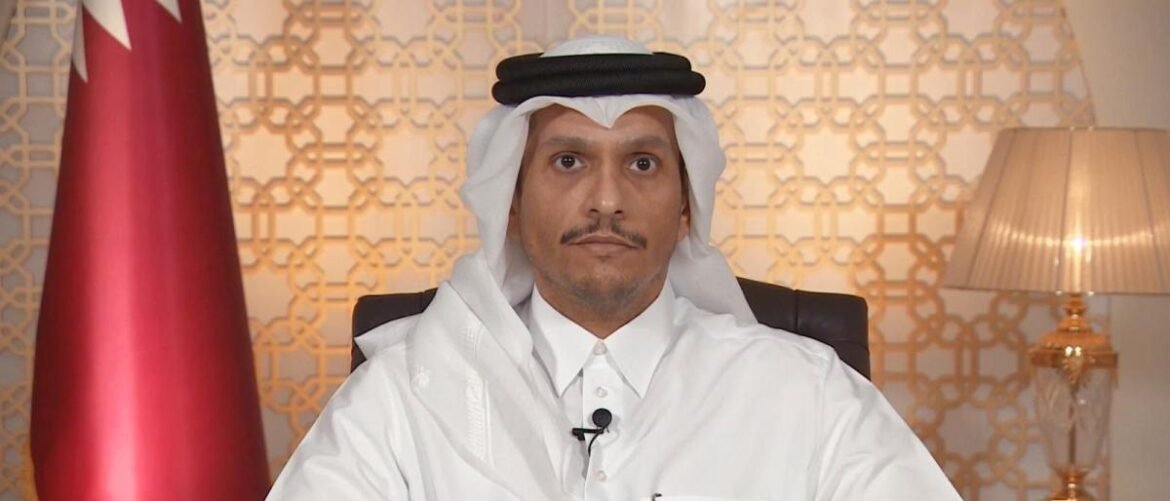 Man in traditional Qatari attire (white thobe and ghutra) sits at a desk with a lapel microphone in front, a red flag to his left and a lamp to the right in a patterned backdrop.
