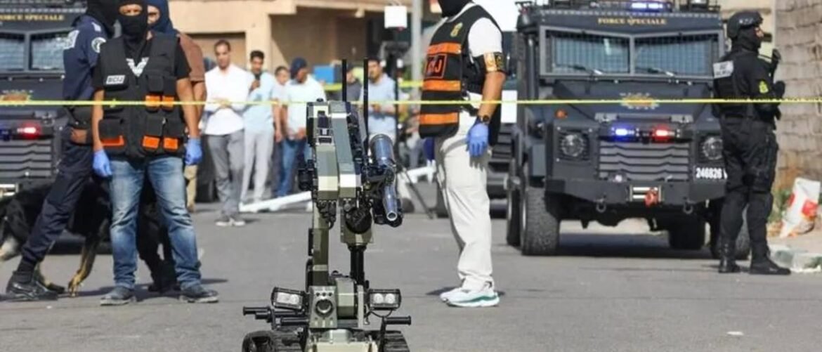 Cordoned street scene with masked officers, armored vehicles, and a wheeled bomb-disposal robot at the center of a security operation.