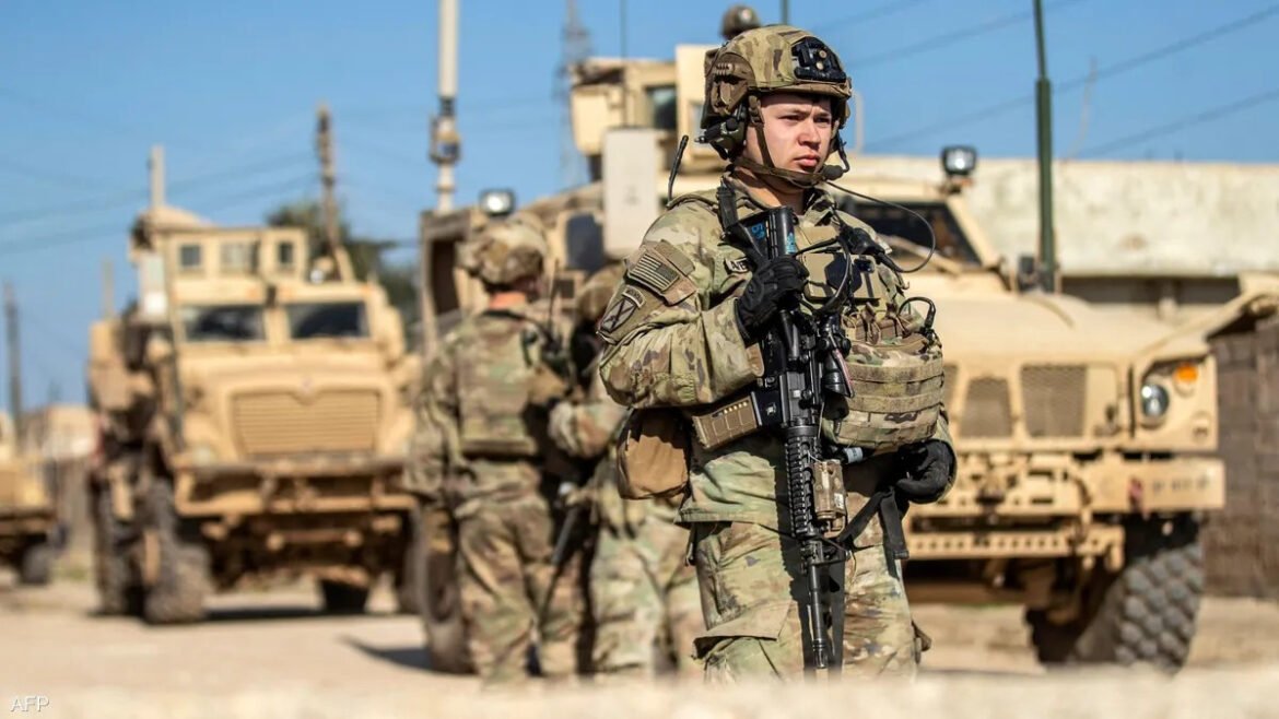 Soldier in desert camouflage with helmet and rifle stands in front of armored vehicles at a base under a blue sky.